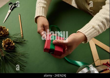 Une femme en gros plan noue le présent, enveloppée de papier rouge avec du ruban vert. Emballage des cadeaux de Noël et du nouvel an. Lendemain de Noël Banque D'Images