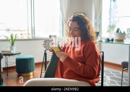 Jeune souriante jolie femme tenant le téléphone boire un café se détendre à la maison. Banque D'Images