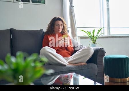Une jeune femme décontractée assise sur un canapé à la maison à l'aide d'un téléphone portable. Banque D'Images