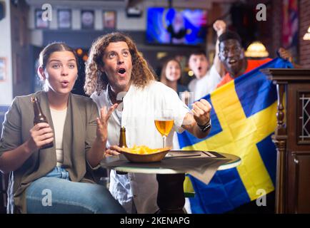 Fans de football émotionnels et variés, drapeau de la Suède, verres de bière et en-cas au bar Banque D'Images