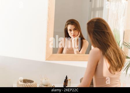 La jeune femme regarde dans le miroir de la salle de bains à la maison le matin. Bouleversé jeune femme malheureuse avec la peau malsaine, concept de traitement. Banque D'Images