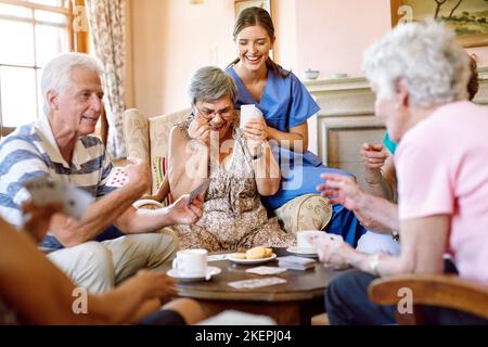 Pour le gagnant aller le butin. Un groupe de personnes âgées heureux jouant un jeu de carte dans leur maison de retraite tandis qu'une infirmière regarde. Banque D'Images