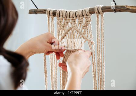 Femme travaillant sur un morceau de macrame suspendu d'un bâton. IL est toujours en cours, seulement partiellement sevrés. Sur un mur blanc. Banque D'Images