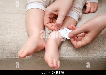 La mère colle un pansement médical adhésif sur la jambe du bébé. Main de maman avec ruban adhésif de protection collant et pied d'enfant. Enfant âgé de un vous Banque D'Images