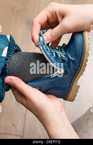 La mère met des chaussures bleues sur le pied de bébé enfant assis dans le couloir de la maison. Femme maman vêtue chaud bottes vêtements sur enfant pour l'hiver marcher dans c Banque D'Images