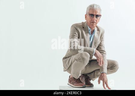 le vieil homme avec des lunettes de soleil étant frais, accroupi et les bras de maintien à la mode posent devant le fond gris Banque D'Images