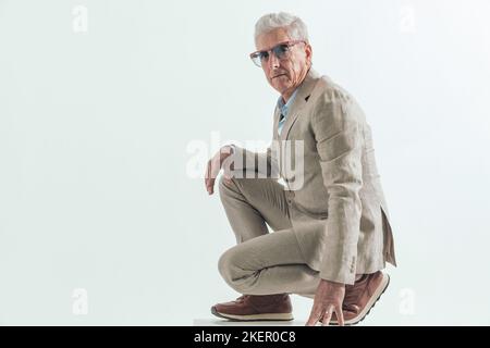 vue latérale du vieil homme dans son 60s portant des lunettes de soleil et accroupi, tenant le bras sur le genou et posant sur fond gris en studio Banque D'Images