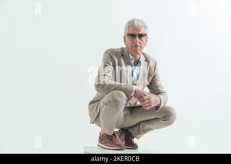 cool mode vieux homme dans ses 60s coudes de maintien sur les genoux et crouching, posant d'une manière sûre sur fond gris Banque D'Images