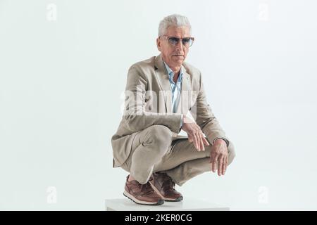 bon look élégant homme en costume accroupi avec les mains sur les genoux, en portant des lunettes de soleil et en posant tout en regardant loin sur fond gris Banque D'Images