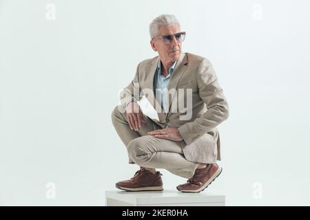 cool homme dans son 60s avec des cheveux gris regardant sur le côté et crouching devant le fond gris en studio Banque D'Images