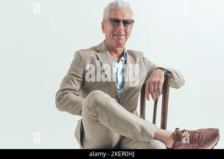 heureux homme élégant dans son 60s souriant, tenant la cheville sur le genou et assis sur une ancienne chaise en bois devant un fond gris Banque D'Images