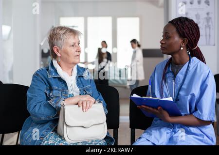 Une infirmière afro-américaine remplit les formalités administratives d'admission des femmes âgées. Médecin notant les symptômes des patients âgés dans le hall de l'hôpital gériatrique. Médecin généraliste interrogeant une femme âgée dans la salle d'attente de l'hôpital. Banque D'Images