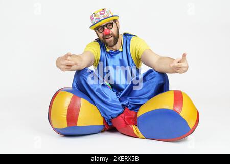 Concept de vacances. Un clown dans un costume bleu et jaune vif, des lunettes et un chapeau est assis sur le sol et des gestes avec ses mains. Banque D'Images