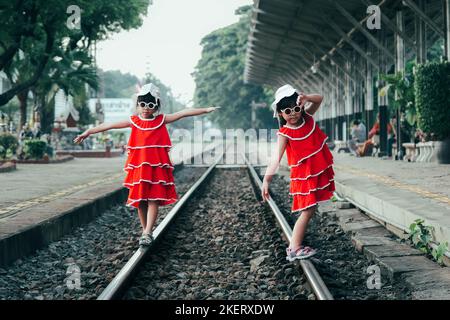 Deux petites sœurs en robe rouge marchent le long des voies de train en été. Les sœurs marchaient sur les voies de la gare. Voyages et transport Banque D'Images