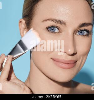 Belle femme appliquant la crème de ton sur son visage parfait avec une brosse cosmétique, isolée sur fond bleu. cosmétiques pour le visage, fond de teint Banque D'Images