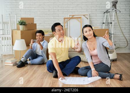 Un couple asiatique enthousiaste assis au sol d'un nouvel appartement et préparant la décoration de son salon spacieux pendant que son fils souriant s'est enveloppé dans un jeu sur smartphone Banque D'Images