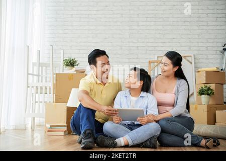 Joyeuse famille de trois s'amuser ensemble tout en s'asseyant sur le sol d'un nouvel appartement spacieux et en partageant des idées concernant la conception de la salle de séjour les uns avec les autres, des tas de boîtes mobiles sur l'arrière-plan Banque D'Images