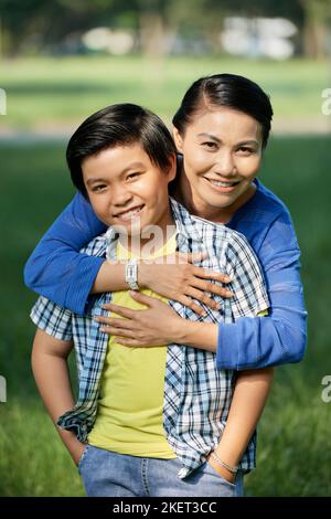 Belle femme d'âge moyen regardant l'appareil photo avec un sourire charmant et embrassant son petit fils tout en profitant de la chaude journée ensoleillée dans le parc public, portrait de groupe Banque D'Images