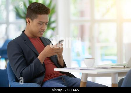 Souriant ouvrier vietnamien blanc de collier assis à la table de café et de texter avec un ami sur le smartphone tout en prenant une pause café, portrait tourné Banque D'Images