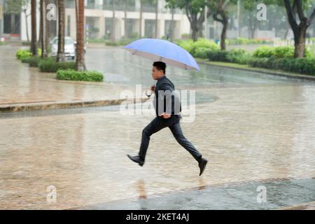 Un jeune homme d'affaires vietnamien portant un costume classique traversant une route tout en se précipitant à la maison après une dure journée de travail dans la pluie battante, il tient un parapluie à la main Banque D'Images