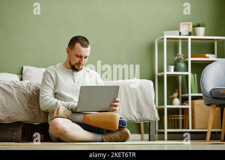 Portrait minimal d'un homme avec une jambe prothétique à l'aide d'un ordinateur portable assis au sol à la maison, espace de copie Banque D'Images