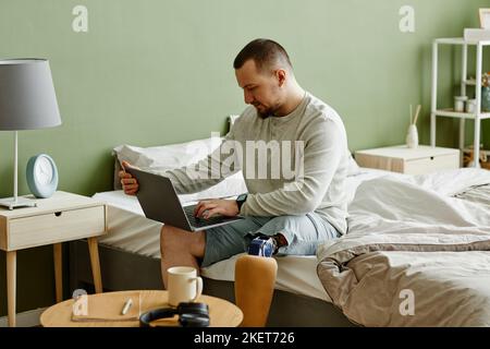 Portrait d'un homme avec une jambe prothétique à l'aide d'un ordinateur portable tout en étant assis sur le lit à la maison, espace de copie Banque D'Images