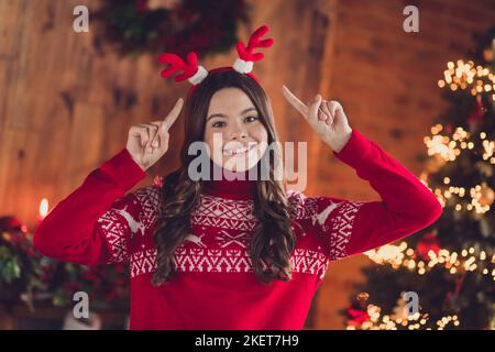 Portrait d'une jeune fille gaie optimiste avec un chandail en forme de cheveux ondulés habillé rouge ornement dirigeant vers le bandeau renne dans la maison à l'intérieur Banque D'Images