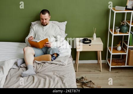 Vue en grand angle sur un jeune homme avec un livre de lecture prothétique sur la jambe au lit tout en se relaxant à la maison, espace de copie Banque D'Images