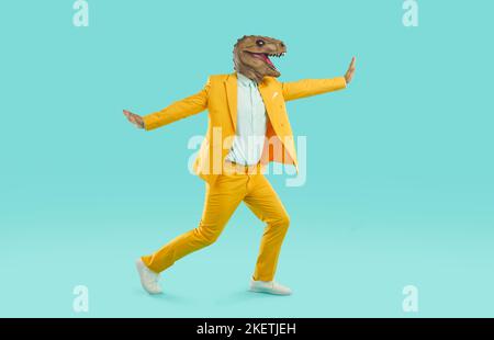 Étrange homme en costume jaune et drôle masque de dinosaure courant et dansant sur fond bleu Banque D'Images