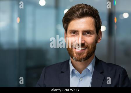 Gros plan photo d'un homme d'affaires mûr avec une barbe regardant la caméra et souriant, un investisseur banquier réussi à l'intérieur d'un immeuble de bureau moderne au travail, un homme en costume de soirée Banque D'Images