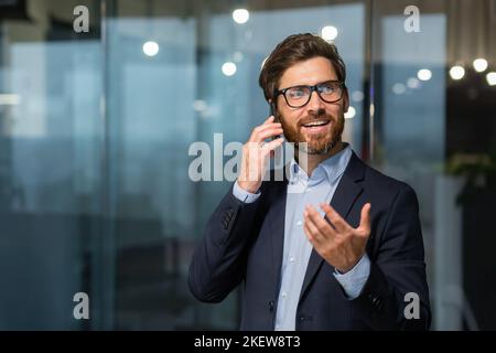 Homme d'affaires mature dans un costume d'affaires parlant au téléphone, le directeur est satisfait du résultat de la réalisation et le travail parle du résultat positif, l'homme est au travail à l'intérieur du bureau. Banque D'Images