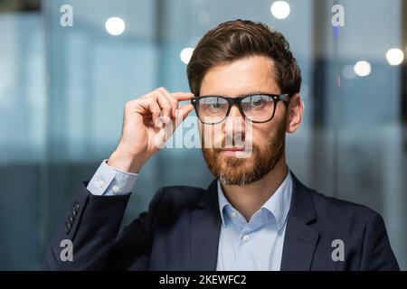 Portrait en gros plan d'un homme d'affaires sérieux et sérieux avec barbe, homme en chemise et lunettes regardant l'appareil photo, startuper travaillant à l'intérieur d'un immeuble de bureau moderne. Banque D'Images