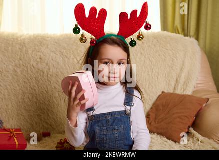 Belle petite fille portant un drôle de cervidés, tenant une boîte cadeau en forme de coeur, assis sur un canapé dans la salle de séjour Banque D'Images