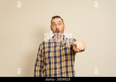 Homme hispanique barbu vêtu d'une chemise à carreaux pointant du doigt surpris devant, expression étonnante avec bouche ouverte, quelque chose devant. Banque D'Images