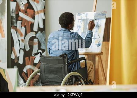 Vue arrière d'un peintre afro-américain peint en fauteuil roulant sur toile tout en étant assis devant un chevalet en studio ou en classe Banque D'Images