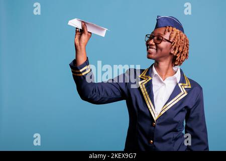 Portrait d'une femme afro-américaine qui tient un avion en papier prêt à être lancé. Hôtesse avec verres ronds et uniforme formel en studio tourné sur fond bleu. Banque D'Images