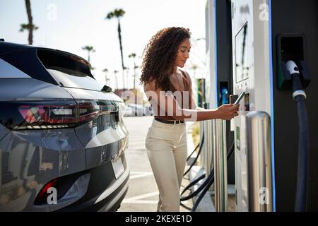 Jeune femme qui balaie un smartphone au poste de charge de véhicule électrique Banque D'Images