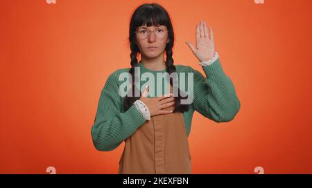 Je jure d'être honnête. Une jeune femme sincère et responsable qui tient la main pour prêter serment, promettant d'être honnête et de dire la vérité, en gardant la main sur la poitrine. Fille latine isolée sur fond orange studio Banque D'Images