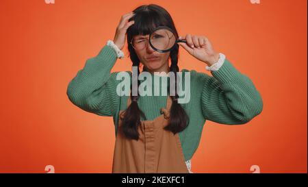 Chercheur scientifique femme tenant la loupe près du visage, regardant dans la caméra avec les grands zooms yeux drôles, la recherche, l'analyse. Jeune fille adulte isolée sur fond orange studio Banque D'Images