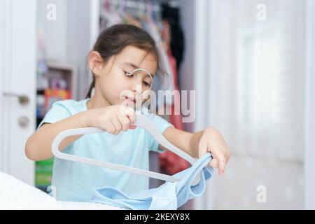 Belle petite fille avec de longs cheveux noirs apporte l'ordre de garde-robe, met tout à sa place dans le placard. Garde-robe avec vêtements pour enfants. Mise au point sélective sur le rack. Banque D'Images