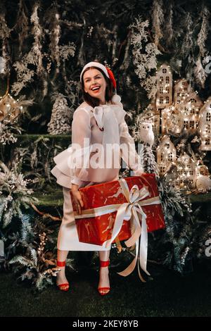 Une fille dans un chapeau de père Noël avec un grand cadeau de Noël dans ses mains sur une île de conte de fées.femme souriante en vêtements blancs sur le fond de Noël Banque D'Images