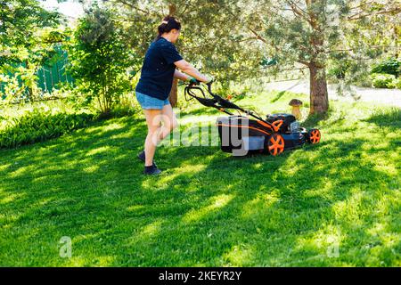 Jardinière féminine aux cheveux bruns vêtue de vêtements décontractés utilisant une tondeuse à essence ou un coupe-herbe dans le jardin. Activité le week-end Banque D'Images