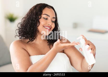 Bonne femme afro-américaine chubby appliquant de la lotion hydratante à la main, dame enveloppée dans une serviette après le bain Banque D'Images