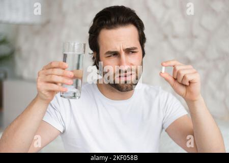 Portrait de triste millénaire homme caucasien avec le chaume est assis sur le lit tient un verre d'eau et de pilule, souffre de douleur Banque D'Images