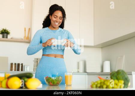 Concept de blogging alimentaire. Blogueur de forme physique noir prenant la photo d'un dîner sain sur le téléphone portable, la cuisine de salade dans la cuisine Banque D'Images