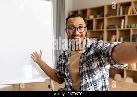 Joyeux tuteur arabe souriant à l'appareil photo et pointant vers le tableau blanc, ayant cours en ligne en classe Banque D'Images