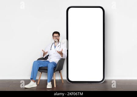 Concept de télémédecine. Beau médecin masculin assis sur la chaise près du smartphone Big Blank Banque D'Images