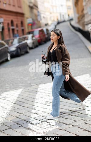 pleine longueur de femme tendance en manteau et jeans tenant des lunettes de soleil en traversant la rue à prague, image de stock Banque D'Images