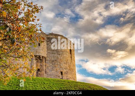 Une ancienne tour située sur un remblai d'herbe. Un arbre d'automne est au premier plan et un ciel nuageux est au-dessus. Banque D'Images