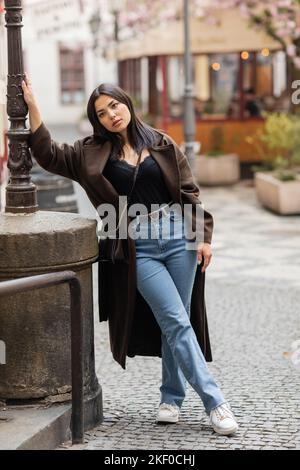 jeune femme brune en manteau brun et jeans penchée sur le lampadaire dans la rue à prague, image de stock Banque D'Images